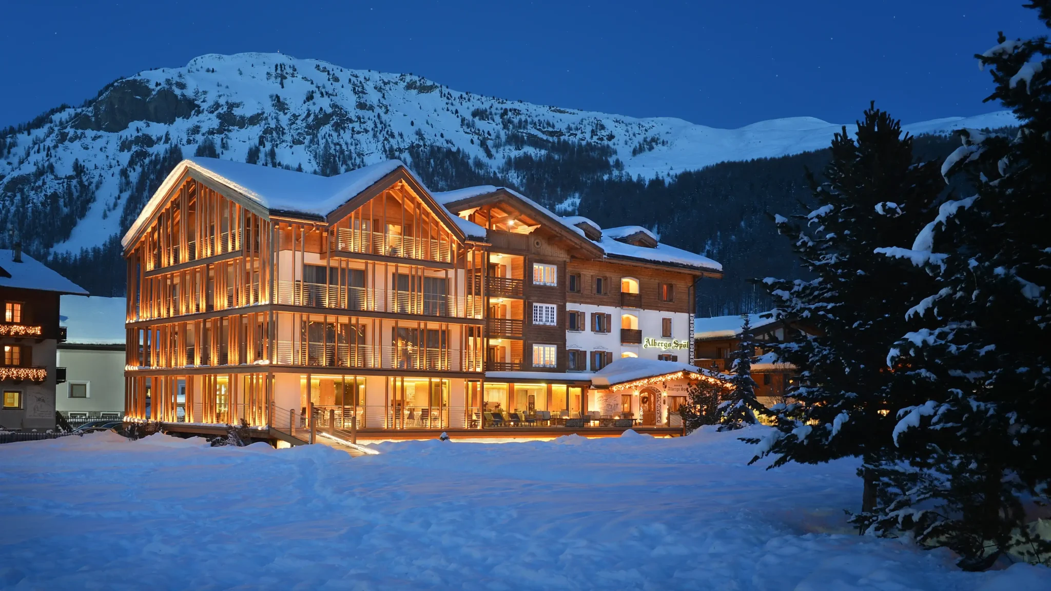Hotel di montagna illuminato di notte con montagna innevata sullo sfondo.