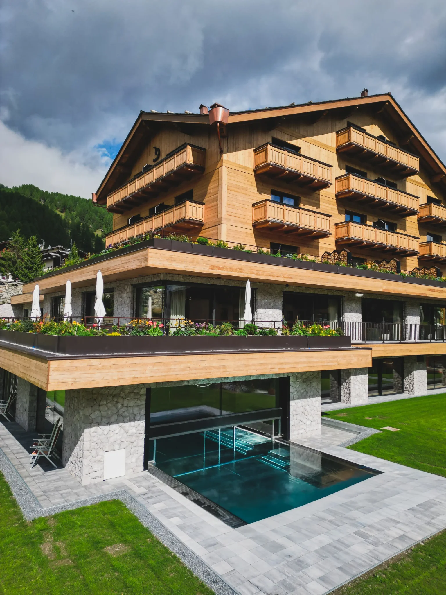 Edificio in legno e pietra con terrazze e piscina su prato verde sotto cielo nuvoloso.