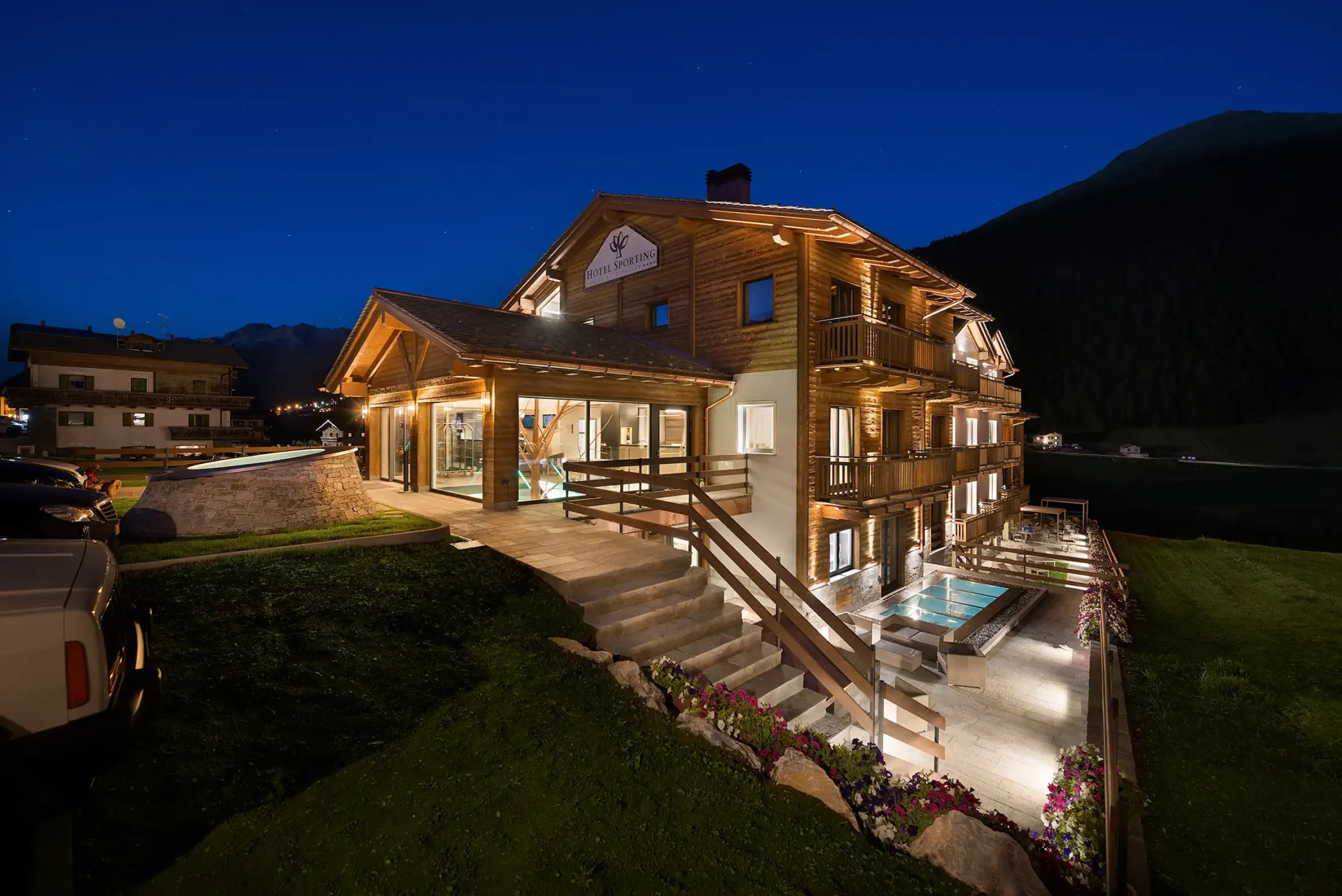 Hotel di montagna in legno illuminato di notte, con piscina esterna e panorama alpino.