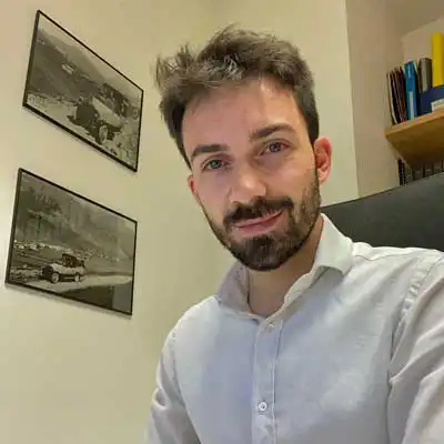 Mi dispiace, non so chi sia la persona nella foto. Descrizione: Uomo con barba in camicia bianca, seduto in un ufficio con due foto storiche sul muro.